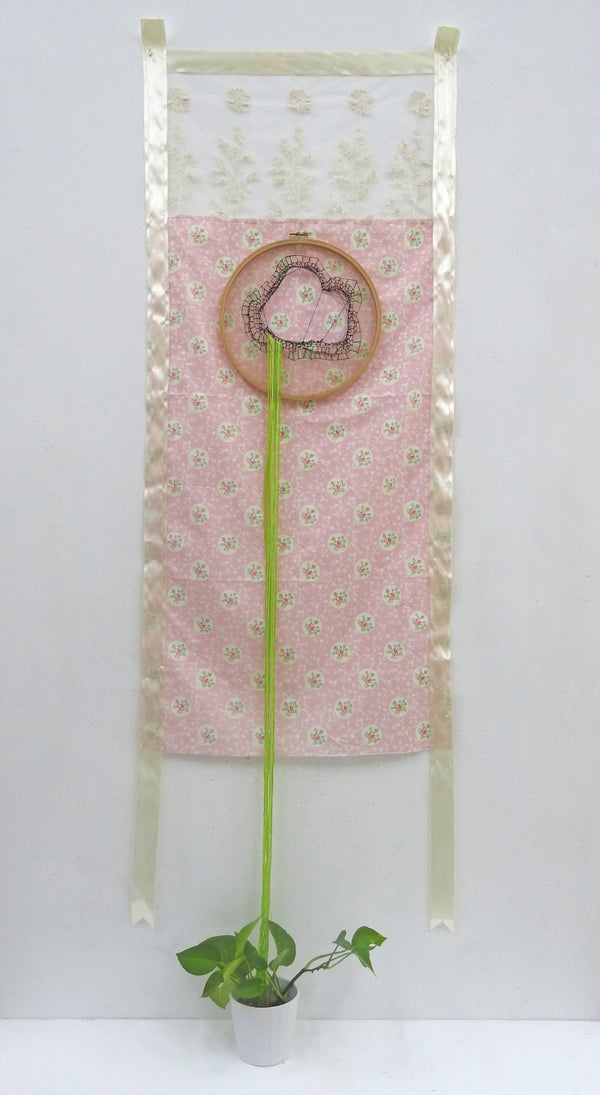 Suture by Wendy Tai is a found object assemblage featuring an embroidery hoop attached on a domestic pink floral fabric, with green fibres hanging from the hoop to a potted plant on the ground. 